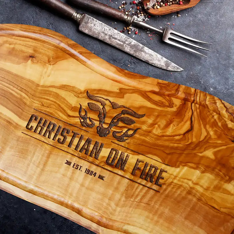 Motiv "On Fire" mit stilisiertem Rinderkopf mit Flammen und Personaliserungsoption mit Name in Großbuchstaben und Datum - Mediterranes Schneidebrett aus Olivenholz mit Saftrille mit und ohne Grifflasche oder Handgriff - Personalisiertes Männergeschenk von Jeske Design
