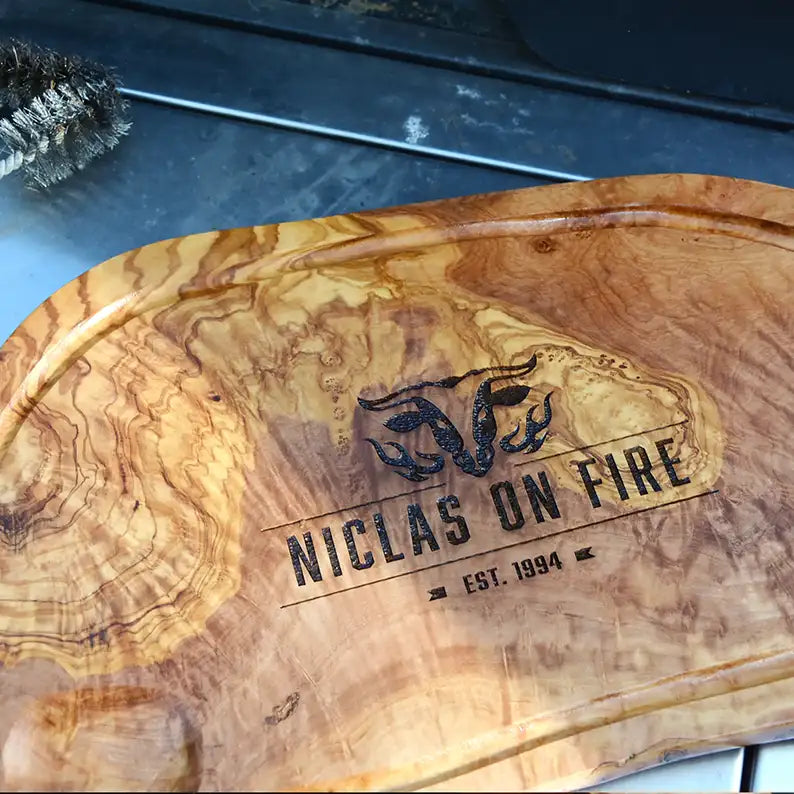 Motiv "On Fire" mit stilisiertem Rinderkopf mit Flammen und Personaliserungsoption mit Name in Großbuchstaben und Datum - Mediterranes Schneidebrett aus Olivenholz mit Saftrille mit und ohne Grifflasche oder Handgriff - Personalisiertes Männergeschenk von Jeske Design