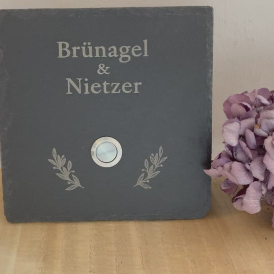 Personalisiertes Klingelschild aus Schiefer mit Edelstahl Taster mit oder ohne LED-Beleuchtung 1. Zeile in kleiner Druckschrift für Familie / Vornamen / Willkommen bei; 2. Zeile mit klassischem Schriftbild in der Größe 15x15 cm, 20x15 cm, 26x16 cm oder 30x20 cm. Hier mit Edelstahltaster matt
