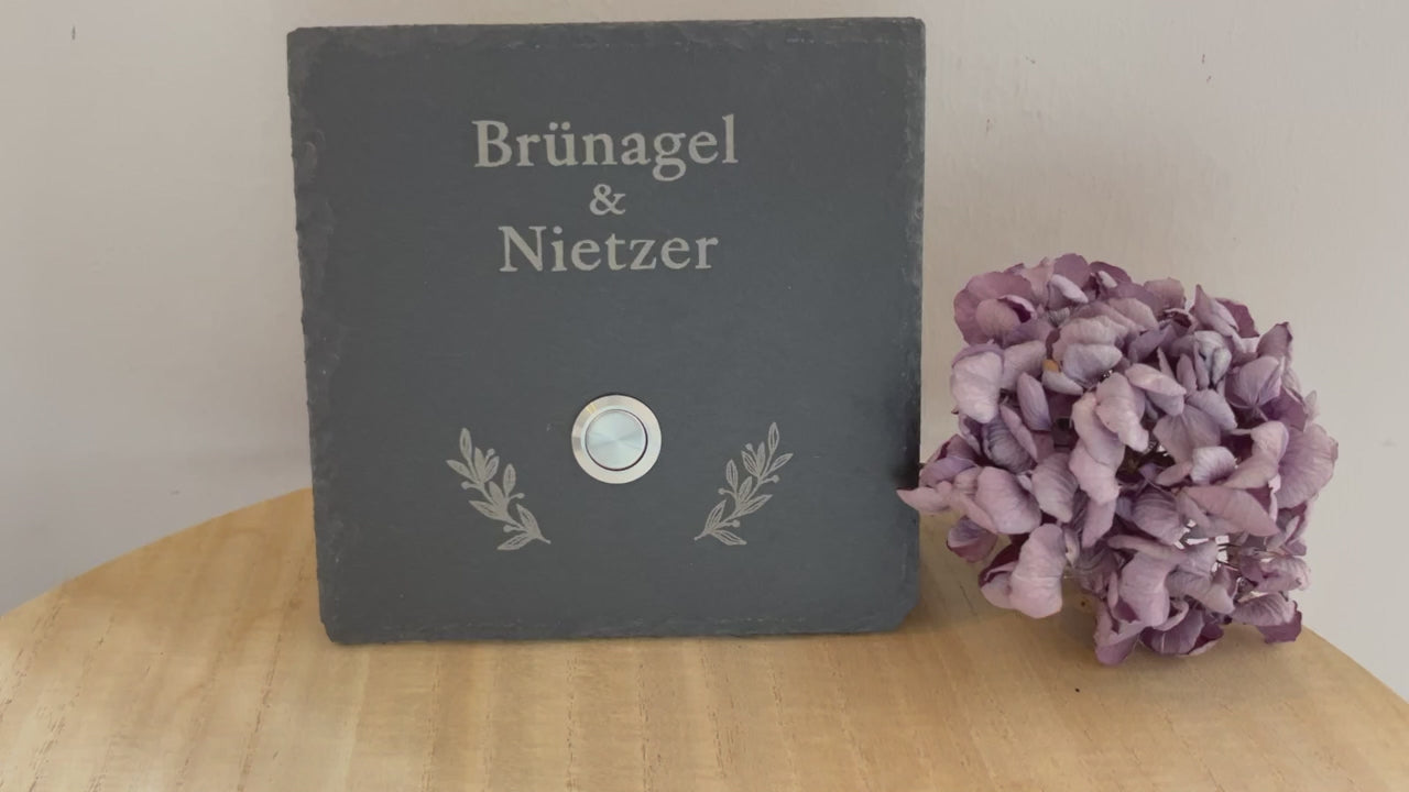 Personalisiertes Klingelschild aus Schiefer mit Edelstahl Taster mit oder ohne LED-Beleuchtung 1. Zeile in kleiner Druckschrift für Familie / Vornamen / Willkommen bei; 2. Zeile mit klassischem Schriftbild in der Größe 15x15 cm, 20x15 cm, 26x16 cm oder 30x20 cm. Hier mit Edelstahltaster matt