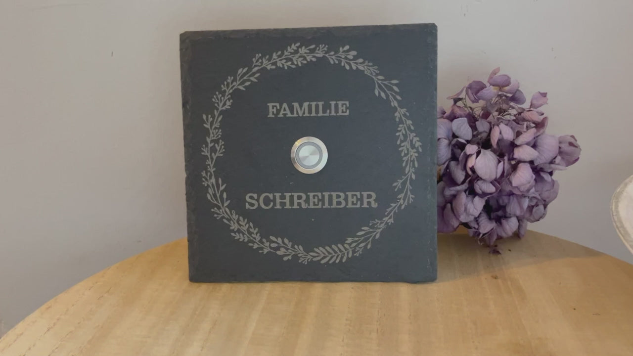 Personalisiertes Klingelschild aus Schiefer mit Edelstahl Taster mit LEDBeleuchtung oder ohne Beleuchtung. Zarter Blätterkranz rund mit Klingelknopf innen. Text in klassischer Druckschrift 2-zeilig oben und unten in der Größe ø 15 cm rund oder 15x15 cm, 20x15 cm, 26x16 cm oder 30x20 cm.